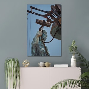 May include: A canvas print featuring a weathered bronze statue of a man holding a tool, positioned beneath a large bell and its supporting structure. The artwork has a blue sky background and is displayed on a wall.