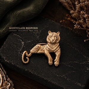 Könnte beinhalten: Goldfarbene Tigerbrosche auf dunkler, strukturierter Oberfläche. Die Brosche weist detaillierte Streifen und Gesichtszüge auf. Der Text "MONTCLAIR BESPOKE LUXURY TAILORED FOR YOU" ist sichtbar. Ein dekoratives Accessoire.