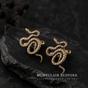 May include: Two gold-coloured cufflinks, each crafted in the form of a detailed snake. They are presented on a dark, textured surface. The text "MONTCLAIR BESPOKE LUXURY TAILORED FOR YOU" is displayed below.