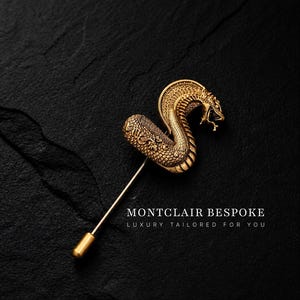 Puede incluir: Broche de solapa dorado con forma de serpiente y detalles intrincados. El broche presenta un diseño de cobra con la boca abierta sobre un fondo oscuro. El texto "MONTCLAIR BESPOKE LUXURY TAILORED FOR YOU" está presente.
