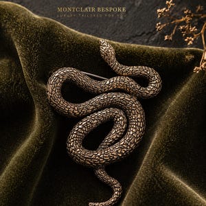 Puede incluir: Broche de serpiente en tono bronce con detalles de escamas, sobre terciopelo verde oscuro. La serpiente está enrollada, con la cabeza levantada. El texto "MONTCLAIR BESPOKE LUXURY TAILORED FOR YOU" es visible.