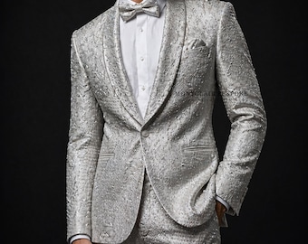 May include: Silver suit with a textured pattern, featuring a matching blazer, trousers, and bow tie. The suit has a shawl collar and a pocket square. The text "MONTCLAIRE BESPOKE" is visible on the jacket.