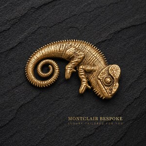May include: A detailed, golden-colored chameleon figurine with a curled tail, resting on a textured black surface. The figurine has intricate detailing and is positioned in a curved pose. The text "MONTCLAIR BESPOKE" is visible.