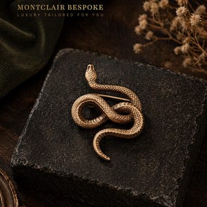 Puede incluir: Broche de serpiente dorada con detalles intrincados, colocada sobre una superficie oscura y texturizada. La broche representa una serpiente enroscada, con un alfiler para sujetarla. El texto "MONTCLAIR BESPOKE LUXURY TAILORED FOR YOU" está presente.