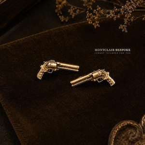 Op de afbeelding: Twee goudkleurige revolvermanchetknopen met gedetailleerde ontwerpen. Ze worden getoond op een donkerbruine achtergrond. De afbeelding bevat de tekst "MONTCLAIR BESPOKE LUXURY TAILORED FOR YOU".