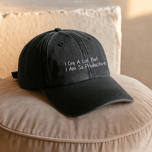 May include: A black baseball cap featuring the white embroidered text I Cry A Lot But I Am So Productive. The cap has a structured crown, a curved brim, and a series of stitched panels meeting at a central button on the top of the head.
