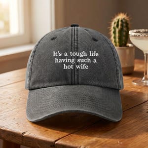 Op de afbeelding: Een grijze baseballpet met de witte tekst "It's a tough life having such a hot wife". De pet heeft een gebogen klep en een stoffen bandsluiting.