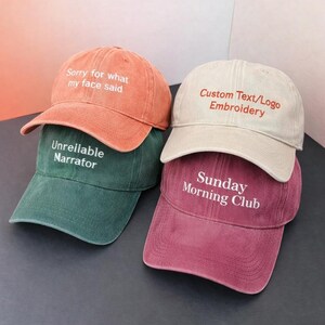 May include: Four baseball caps in shades of orange, green, beige, and burgundy are stacked. Each cap features embroidered text: "Sorry for what my face said," "Unreliable Narrator," "Custom Text/Logo Embroidery," and "Sunday Morning Club."