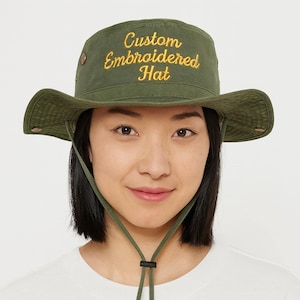 Puede incluir: Sombrero de pescador verde oliva con el texto "Custom Embroidered Hat" en escritura amarilla. El sombrero tiene un ala ancha y una correa para la barbilla. La persona lleva el sombrero.