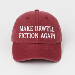 May include: A maroon baseball cap with the phrase "MAKE ORWELL FICTION AGAIN" embroidered in white. The cap has a curved brim and a button on top. The hat is a solid color and appears to be made of cotton.
