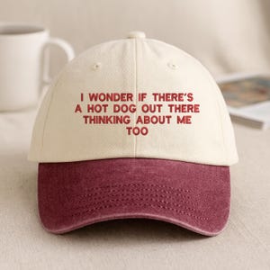 Può includere: Un cappellino da baseball con corona color crema e visiera bordeaux. Il cappellino presenta la scritta "I WONDER IF THERE'S A HOT DOG OUT THERE THINKING ABOUT ME TOO" in lettere bordeaux. Un accessorio casual.
