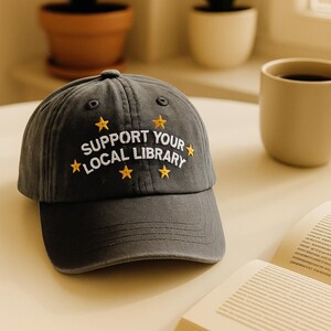 May include: A dark gray baseball cap with the embroidered text "SUPPORT YOUR LOCAL LIBRARY" in white and yellow star accents. The cap is made of a soft fabric and has a curved brim.