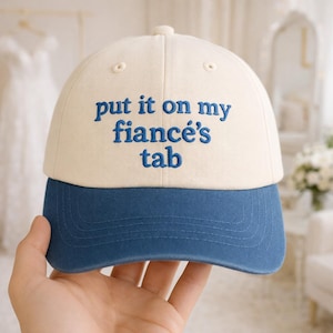 May include: A cream and blue baseball cap with the embroidered text "put it on my fiancé's tab". The cap features a curved peak and a button on top. The text is in blue, and the cap is a stylish accessory.