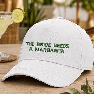 Może przedstawiać: Biała czapka z daszkiem z wyhaftowanym zielonym napisem THE BRIDE NEEDS A MARGARITA na przednim panelu. Czapka posiada usztywnianą koronę, zakrzywiony daszek oraz guzik na górze, wykonana jest z jednolitego białego materiału z zielonymi przeszyciami.
