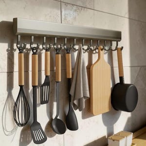 Op de afbeelding: Een aan de muur gemonteerd metalen keukenrek met twaalf haken. Aan de haken hangen een garde, twee sleufspatels, een soeplepel, een dichte spatel, een grijze doek, een houten snijplank en een kleine zwarte steelpan.