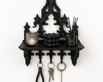 May include: A black wall-mounted shelf with an ornate, pointed arch backboard. The shelf holds a ribbed glass candle holder, a black bubble candle, and a black reed diffuser bottle labeled Noir. Three metal hooks hang below with keychains.