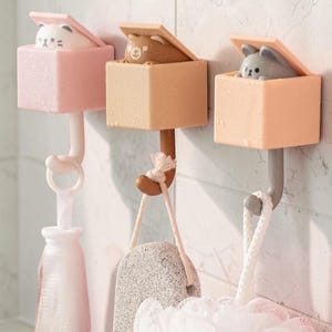 Puede incluir: Tres ganchos de pared decorativos en colores pastel, cada uno con un animal diferente asomando por una caja. Los ganchos están diseñados para colgar artículos como accesorios de baño. El primer gancho es rosa con un gato, el segundo es marrón con un oso y el tercero es melocotón con un conejo.