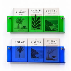 May include: Two horizontal wall-mounted shelves made of translucent acrylic. The top shelf is vibrant green, holding books titled The Kinfolk Home, Matisse The Cut-Outs, and Cereal. The bottom shelf is deep blue, holding Loewe, The Kinfolk Travel, and At Home.