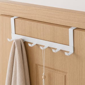 Peut inclure: Un porte-manteau blanc à accrocher sur la porte avec cinq crochets. Une serviette beige est suspendue à l'un des crochets. Le porte-manteau est monté sur une porte en bois de couleur claire. Une ficelle décorative avec une perle en bois est suspendue à un autre crochet.