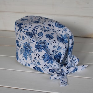 May include: A scrub cap featuring a white base with a detailed blue floral pattern. The head covering has a rounded top section and includes fabric ties at the back, allowing for an adjustable fit. The design displays intricate botanical motifs.