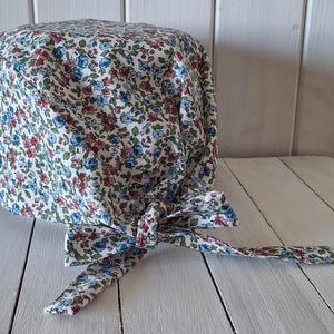 May include: A scrub cap featuring a dense floral pattern with small blue and red flowers on a white background. The cap has a rounded top, a wide band, and two fabric ties at the back that are knotted together to secure the fit around the head.