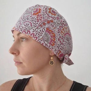 Può includere: Un copricapo aderente caratterizzato da una base bianca con un motivo floreale e paisley ripetuto nei toni del rosa, dell'arancione e del grigio scuro. Il tessuto è annodato sul retro con un piccolo nodo per una vestibilità sicura.