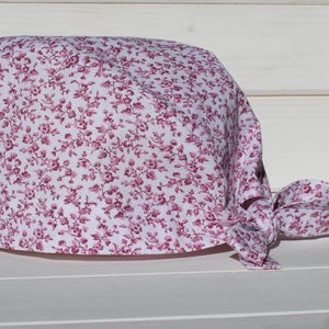 Op de afbeelding: Een operatiemuts van witte stof met een dicht, herhalend patroon van kleine magenta bloemtakjes. De muts heeft een afgeronde bovenkant en is voorzien van twee stoffen linten aan de achterzijde die worden geknoopt voor een goede pasvorm.
