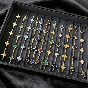 Pode incluir: Exibição de pulseiras douradas e prateadas com pingentes em forma de trevo em várias cores, incluindo verde, azul e preto. As pulseiras estão dispostas numa bandeja preta.