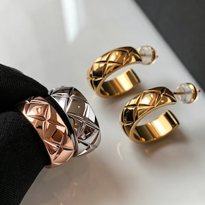 Könnte beinhalten: Ein Paar goldene Creolen und drei Ringe. Die Ohrringe und Ringe haben ein Steppmuster. Ein Ring ist roségold, einer silberfarben und die Ohrringe und der andere Ring sind goldfarben. Der Schmuck liegt auf einer weißen Oberfläche.