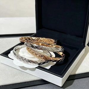 Puede incluir: Cuatro brazaletes con diseño de diamantes en tonos dorados y plateados se exhiben dentro de una caja de joyería blanca con interior negro. Los brazaletes están colocados sobre un cojín blanco en forma de flor.