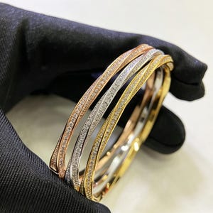 Puede incluir: Tres elegantes brazaletes en oro rosa, plata y oro, cada uno adornado con piedras preciosas brillantes. Los brazaletes se muestran sobre un fondo negro, resaltando su intrincado diseño y su atractivo lujoso. Un sofisticado conjunto de joyas.