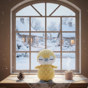 Op de afbeelding: Een handgemaakte gele gehaakte pinguïnpop met gesloten ogen en grijze voeten. Een dennenappel en een mok warme drank staan op een tafel voor een raam met uitzicht op een besneeuwd dorp. De scène roept een gezellige wintersfeer op.