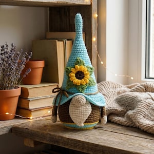 Op de afbeelding: Een handgemaakte gehaakte kabouterpop met een blauwe hoed versierd met een zonnebloem. De kabouter heeft een bruin lichaam, een crème baard en kleine armen en voeten. Het wordt tentoongesteld op een houten plank met boeken en een potplant.