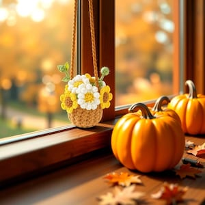 Op de afbeelding: Een gehaakte mand gevuld met gele en witte bloemen hangt bij een raam. Twee oranje pompoenen staan op een houten oppervlak met gevallen bladeren. De scène roept de herfst en seizoensgebonden decoratie op.