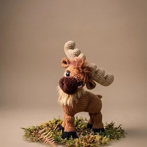 Op de afbeelding: Een gehaakte elandfiguur met een bruin lichaam, een donkerbruine snuit en beige geweien. De eland heeft een pluizige manen en staat op een bedje van groen mos. De ogen zijn groot en expressief.