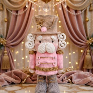 Op de afbeelding: Een handgemaakte gehaakte notenkrakerpop in roze, beige en grijs. De pop heeft een hoge hoed, krullend haar en een gedetailleerd uniform met gouden accenten. De pop staat rechtop en toont zijn ingewikkelde ontwerp en vakmanschap.
