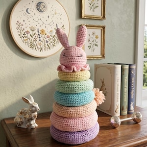Op de afbeelding: Een pastelkleurig gehaakt konijn stapelspeelgoed met een roze konijnenkop en zeven gekleurde ringen. Het speelgoed staat op een houten oppervlak, met een decoratief konijnenbeeldje en ingelijste botanische prenten op de achtergrond.