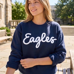May include: A navy blue crewneck sweatshirt featuring the word Eagles written in white, textured, cursive lettering across the chest. The garment has long sleeves with ribbed cuffs and a classic round neckline, providing a simple and clean design.