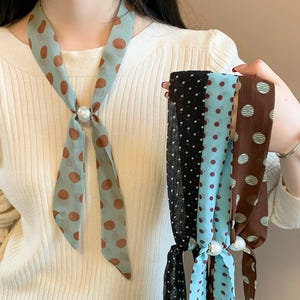 May include: A collection of silk scarves with polka dot patterns. One scarf is light blue with brown dots, fastened with a pearl. Other scarves are black, blue, and brown with white or brown dots. The scarves are tied together with a pearl.