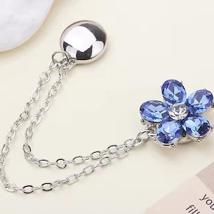 May include: Silver brooch with a flower design, featuring blue gemstone petals and a central clear stone. It's linked to a silver chain and a round, silver backing. The text "YYXaccessory" is visible.