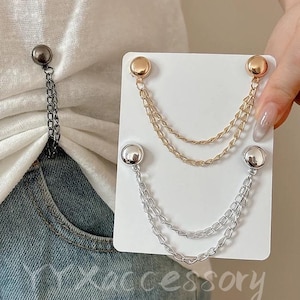 May include: A collection of decorative chain brooches. One brooch is attached to a white shirt and denim jeans. The other two brooches are displayed on a white card. One brooch is gold-toned, and the other is silver-toned. The brooches have a chain design.