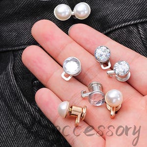 May include: A selection of decorative buttons featuring pearl, crystal, and glitter designs. The buttons are in silver and gold colours, with a small hook for attachment. The word "accessory" is visible in the image.