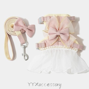 Puede incluir: Conjunto de arnés y correa para perro en rosa claro y crema. El arnés presenta un lazo grande, una falda blanca y detalles de perlas. La correa a juego tiene un lazo y un cierre plateado. El texto "YYXaccessory" está en la parte inferior.