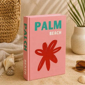 May include: A pink book with the words "PALM BEACH" in teal and a red flower design. The book is on a sandy surface with sunglasses, a seashell, and a woven bag in the background.