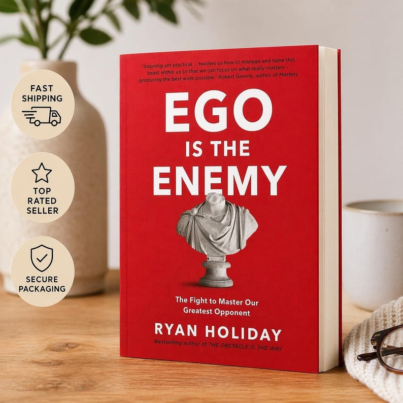 May include: A hardcover book titled "Ego Is The Enemy" by Ryan Holiday. The book has a red cover with white text and a bust illustration. The book is on a wooden surface with a white vase and a mug in the background.