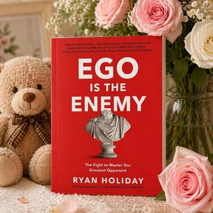 May include: A hardcover book titled "Ego Is The Enemy" with a red cover and white text. The book features a bust illustration. A teddy bear and a bouquet of pink roses are in the background.