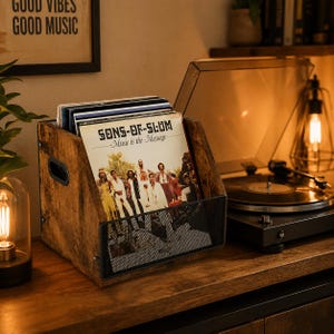 Può includere: Un portadischi in legno con un frontale in rete metallica nera, pieno di dischi in vinile. La copertina dell'album visibile recita "SONS-OF-SLUM - Music is the Message". Il supporto è appoggiato su una superficie di legno, con un giradischi e una lampada vintage sullo sfondo.