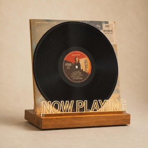 May include: A black vinyl record in its album cover, displayed on a wooden stand with the words "NOW PLAYING" in gold lettering. The album cover is visible behind the record.