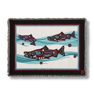 May include: A woven throw blanket with a black border and a white background featuring three stylized salmon swimming in blue water. The salmon are red, blue, and black with geometric patterns.
