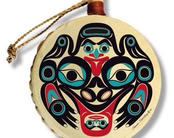 May include: A round, hand-crafted drum with a leather head and a rope handle. The drum is decorated with a colorful, stylized frog design in the style of Northwest Coast Indigenous art. The artist's signature, "Susan  Sewid-Sayers 09", is visible in the lower right corner.